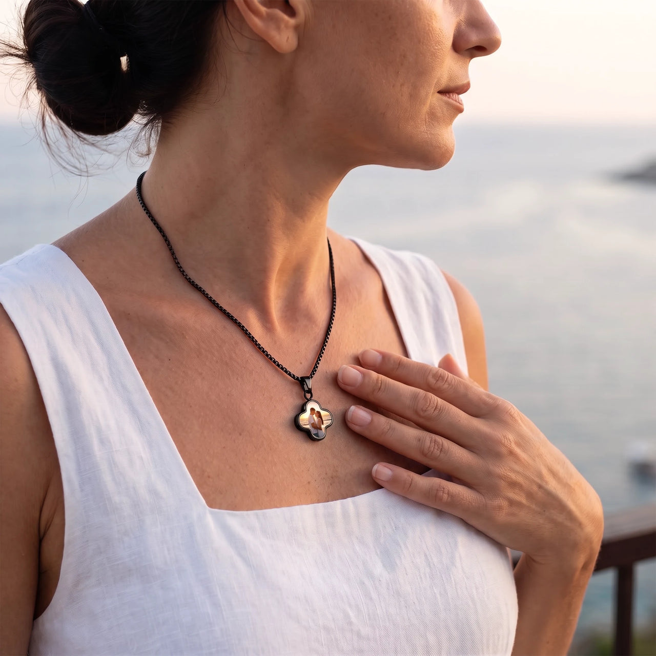 Woman in a white top by the sea, hand on her heart, wearing the black secret clover photo necklace | SKU: TPH-CLVN-BLACK